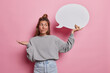 Hesitant woman shrugs shoulders with bewilderment holds white communication bubble cannot make decision dressed in casual grey jumper jeans over against pink background. Unsure questioned female model