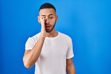 Wall Mural - Young hispanic man standing over blue background hand on mouth telling secret rumor, whispering malicious talk conversation