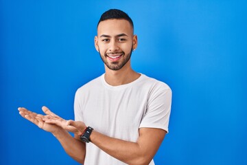 Poster - Young hispanic man standing over blue background pointing aside with hands open palms showing copy space, presenting advertisement smiling excited happy