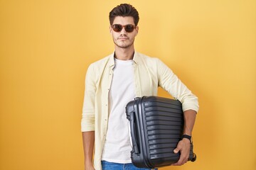 Wall Mural - Young hispanic man holding suitcase going on summer vacation relaxed with serious expression on face. simple and natural looking at the camera.