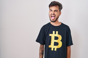 Poster - Young hispanic man with tattoos wearing bitcoin t shirt sticking tongue out happy with funny expression. emotion concept.