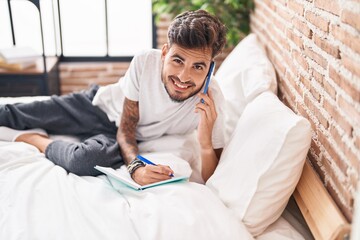 Sticker - Young hispanic man talking on smartphone writing on book at bedroom