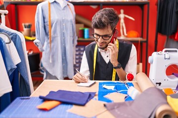 Poster - Young hispanic man tailor talking on smartphone writing on notebook at atelier