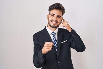 Poster - Young hispanic man with tattoos wearing business suit and tie smiling doing talking on the telephone gesture and pointing to you. call me.