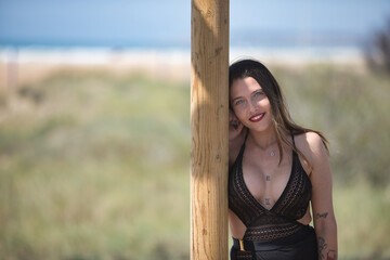 Young and attractive woman, blue eyes, wearing black swimsuit and sarong, leaning on a wooden pole, happy and relaxed. Concept peace, tranquility, relaxation.