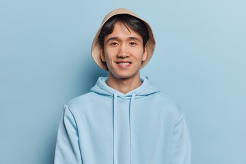 Wall Mural - Portrait of handsome brunet young Asian man smiles gently wears panama and casual hoodie looks satisfied at camera isolated over blue background. People ethnicity and positive emotions concept