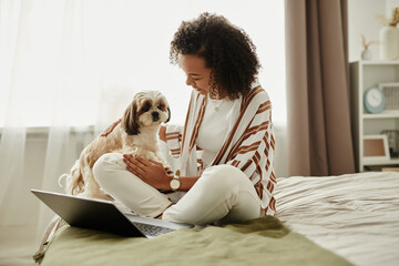 Wall Mural - Candid portrait of young black girl relaxing at home sitting on bed with cute pet dog and using laptop, copy space