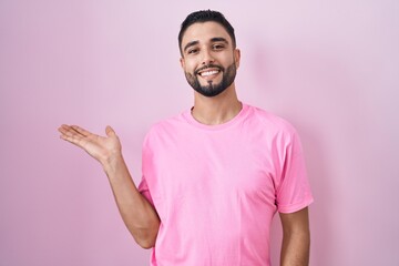 Wall Mural - Hispanic young man standing over pink background smiling cheerful presenting and pointing with palm of hand looking at the camera.