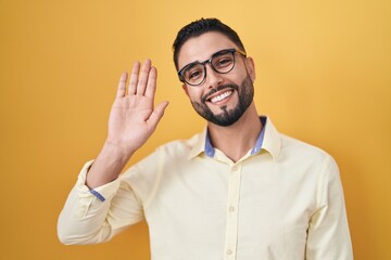 Canvas Print - Hispanic young man wearing business clothes and glasses waiving saying hello happy and smiling, friendly welcome gesture