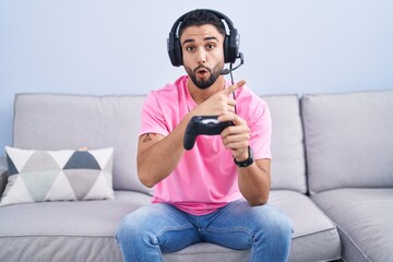 Canvas Print - Hispanic young man playing video game holding controller sitting on the sofa surprised pointing with finger to the side, open mouth amazed expression.