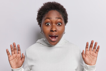 Wall Mural - Attractive oversized dark skinned woman raises palms drops jaw from amazement being stuck in awkward situation holds breath dressed inn sweatshirt isolated over white background stares at horror movie