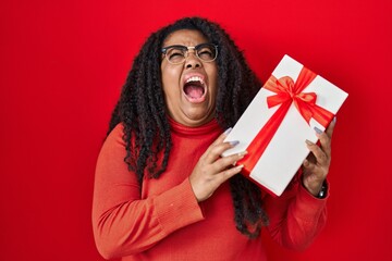 Canvas Print - Plus size hispanic woman holding gift angry and mad screaming frustrated and furious, shouting with anger looking up.