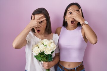 Wall Mural - Hispanic mother and daughter holding bouquet of white flowers peeking in shock covering face and eyes with hand, looking through fingers with embarrassed expression.