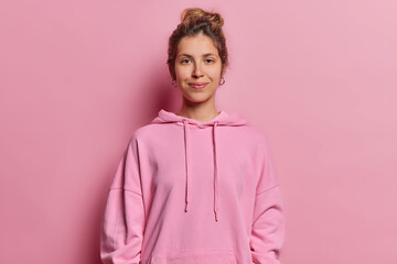 Wall Mural - Portrait of calm pleased woman with dark gathered hair smiles pleasantly looks directly at camera wears casual hoodie isolated over pink background wears piercing and earrings. Monochrome shot