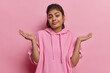 Clueless indecisive Indian woman spreads palm shrugs shoulders looks clueless at camera dressed in casual sweatshirt cannot decide or answer your question isolated over pink background faces dilemma
