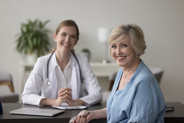 Optimistic, happy mature woman visiting private clinic. Portrait of senior lady patient and female GP therapist on background, smile look at camera. Elder-care, geriatrics, professional consultation