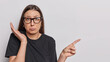Horizontal shot of frustrated woman purses lips keeps palm raised points index finger aside on blank space tells bad news shows mock up space for your advertisement wears spectacles and black t shirt