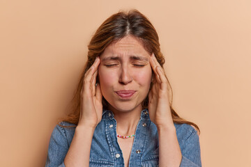 Wall Mural - Frustrated young European woman suffers from headache keeps hands on temples to reveal pain got things messed up tries to calm down during panic wears denim shirt isolated over brown background