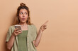 © wayhome.studio  - Horizontal shot of surprised young woman gestures actively points index finger aside on blank space holds mobile phone shows mockup place for your advertisement isolated over brown background