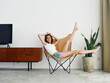 © SHOTPRIME STUDIO - Woman lying in a leather armchair raised arms and legs cheerful happiness and a smile, relaxing at home stylish modern interior with white walls, copy space.