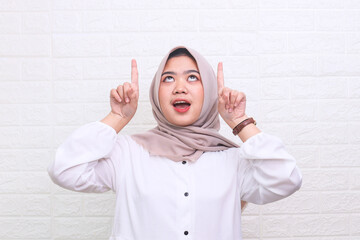 Happy excited Asian Muslim woman in hijab pointing up at copy space with two hands. Cheerful Muslim woman wearing headscarf indicating free place for text above her head 