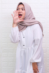 Young beautiful Asian Muslim woman wearing a headscarf shouting and screaming loud with a hand on her mouth. 