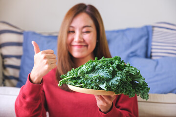 Wall Mural - Portrait image of a young woman making thumb up hand sign while holding a plate of kale leaves