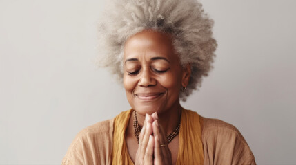 An afro senior woman in a serene moment of prayer, her hands joined and smile radiant on a white studio backdrop. Generative AI