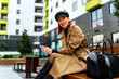 © DusanJelicic - A modern young woman is sitting on a bench and using her phone, she is elegantly dressed in a coat and leather boots