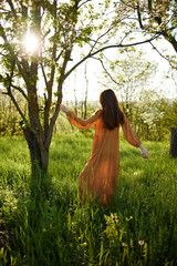 a beautiful, joyful woman stands in a long orange dress, in the countryside, near a tree blooming with white flowers, during sunset, illuminated from the back and reaching for the branches of the tree