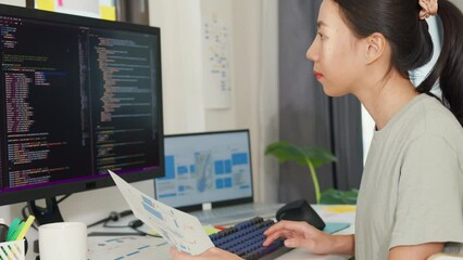 Side view of professional young Asia girl IT development programmer typing on keyboard coding programming fixing data code on computer screen and laptop on table in workroom at house office.