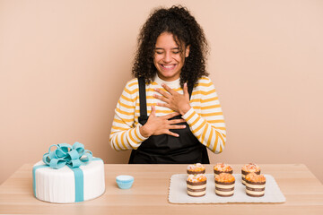 Young african american woman preparing a sweet cake and muffins on a table laughs happily and has fun keeping hands on stomach.