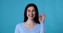 Attractive Caucasian Woman Wearing Blouse Stands On A Blue Background, Gesturing With A Smile And Showing The 'ok' Hand Sign. She Looking Directly At The Camera With A Joyful And Excited Expression.