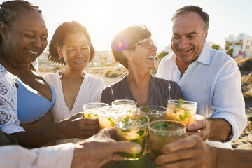 Wall Mural - Senior multiracial friends cheering with alcohol free mojitos on the beach at sunset - Mature people having fun during summer vacation - Focus on right people faces