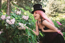 Beautiful Woman With Pink Hair, In A Top Hat And In An Evening Black Dress With A Corset, Standing In The Garden In Azalea Flowers And Laughing