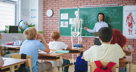 Handsome Caucasian young male teacher of anatomy or biology explaining human body and bones on scelet model to pupils. At school. Education concept. Indoor. Interesting knowledge.