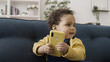 © Synthex - Happy and content African American baby sitting on sofa with smartphone, looking around