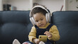 © Synthex - Cute toddler wearing large headphones, sitting on sofa and playing on smartphone
