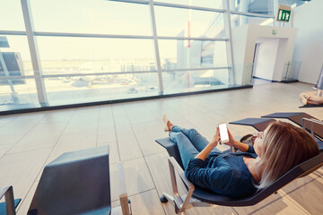 Wall Mural - Travel and technology. Pretty young woman using smartphone while sitting at airport terminal waiting for boarding.