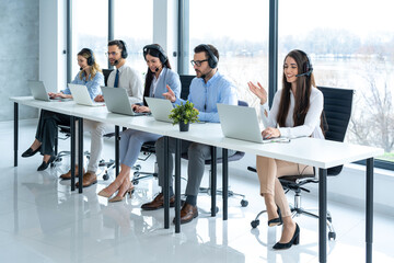 Wall Mural - Long table with customer support operators wearing headsets and using laptop while diligently working at the call center.