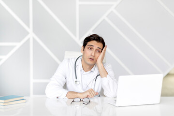 Close up portrait of tired male doctor sitting at the desktop and working on laptop in the office of modern clinic	