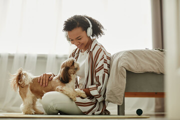 Wall Mural - Side view portrait of young black woman playing with little dog on floor and listening to music, copy space