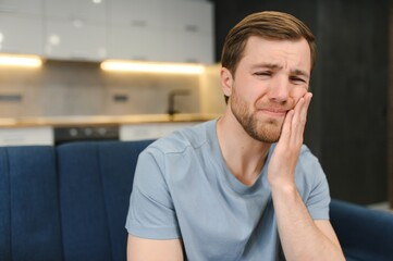 Toothache. Unhappy upset caucasian sad man sits on the couch at home, holds his hand near his cheek, grimaces, has an acute toothache, needs a consultation with a dentist doctor