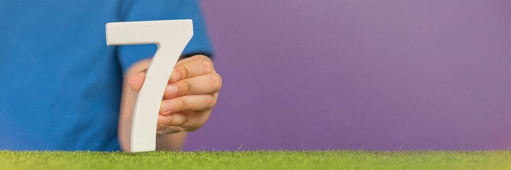 Fototapete - Number 7 in hand. A hand holds a white number seven on green grass and a purple background, seven percent per annum. World Health Day. April 7th. Holiday concept.