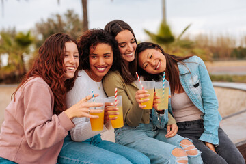 Wall Mural - Portrait of four diverse young women holding drinks while sitting together on beach.