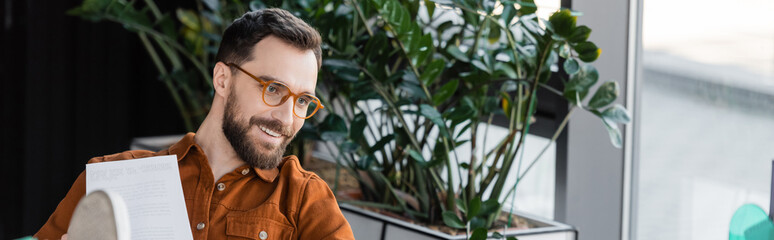 brunette bearded businessman in fashionable eyeglasses and shirt smiling near document and green decorative plants in contemporary office on blurred background, banner