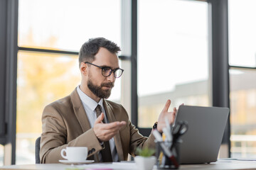 bearded businessman in eyeglasses, stylish blazer and tie gesturing during video conference on laptop near coffee cup and blurred stationery in office