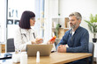 © sofiko14 - Female doctor providing expert check-up and giving medicine to middle-aged patient in hospital office. Focused man listening attentively to recommendations regarding medical history kept on laptop.