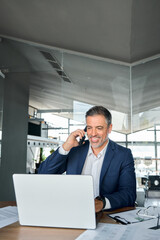 Happy older business man talking on cell phone using laptop in modern office. Mid aged businessman calling on mobile looking at computer discussing digital investment, managing work tasks. Vertical