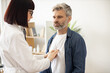 Close up view of serious caucasian man in everyday wear visiting family doctor for full examination in general practice. Young woman in glasses and lab coat checking lungs and heart via stethoscope.
